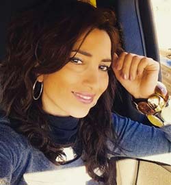 A smiling woman with wavy brown hair, wearing hoop earrings and a bracelet, posing in a car.