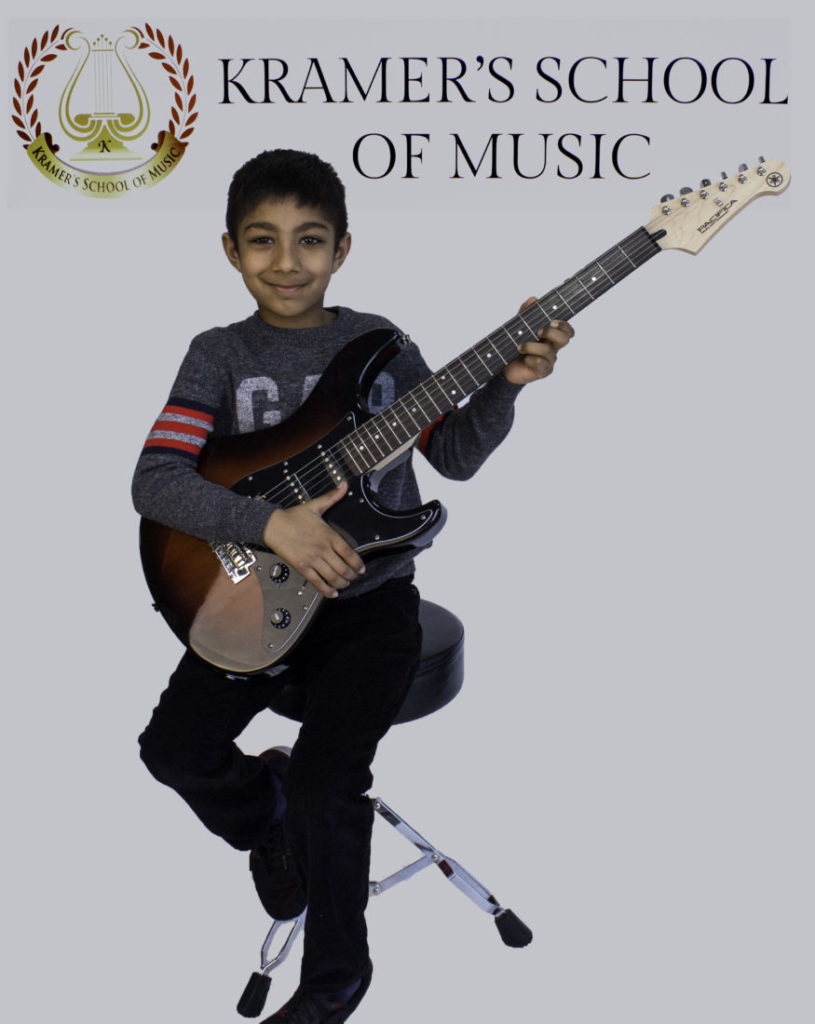 A smiling young boy sits on a stool holding an electric guitar against a light background.