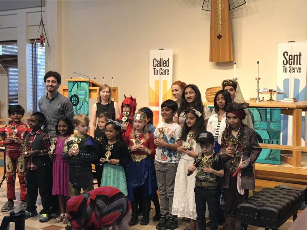 A group of children in costumes hold awards, posing with teachers in a decorated room.