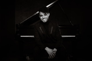 A man in a black outfit sits in front of a grand piano, smiling in a moody black-and-white portrait.